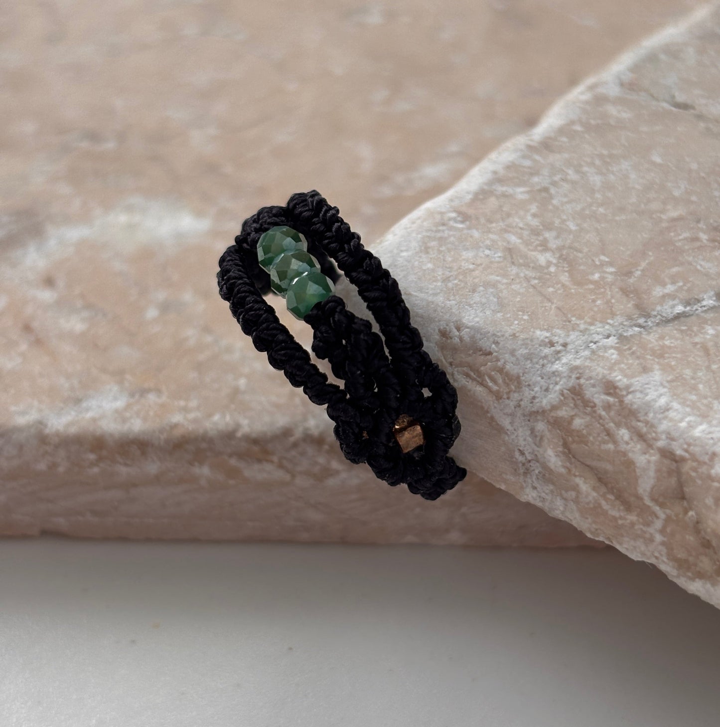A side view of a handmade macrame band ring in black flat waxed nylon cord with a double-wrap braided design and three faceted mint green glass crystal beads with a square gold-tone seed bead accent visible on the underside, leaning against a pink stone surface against a white background,