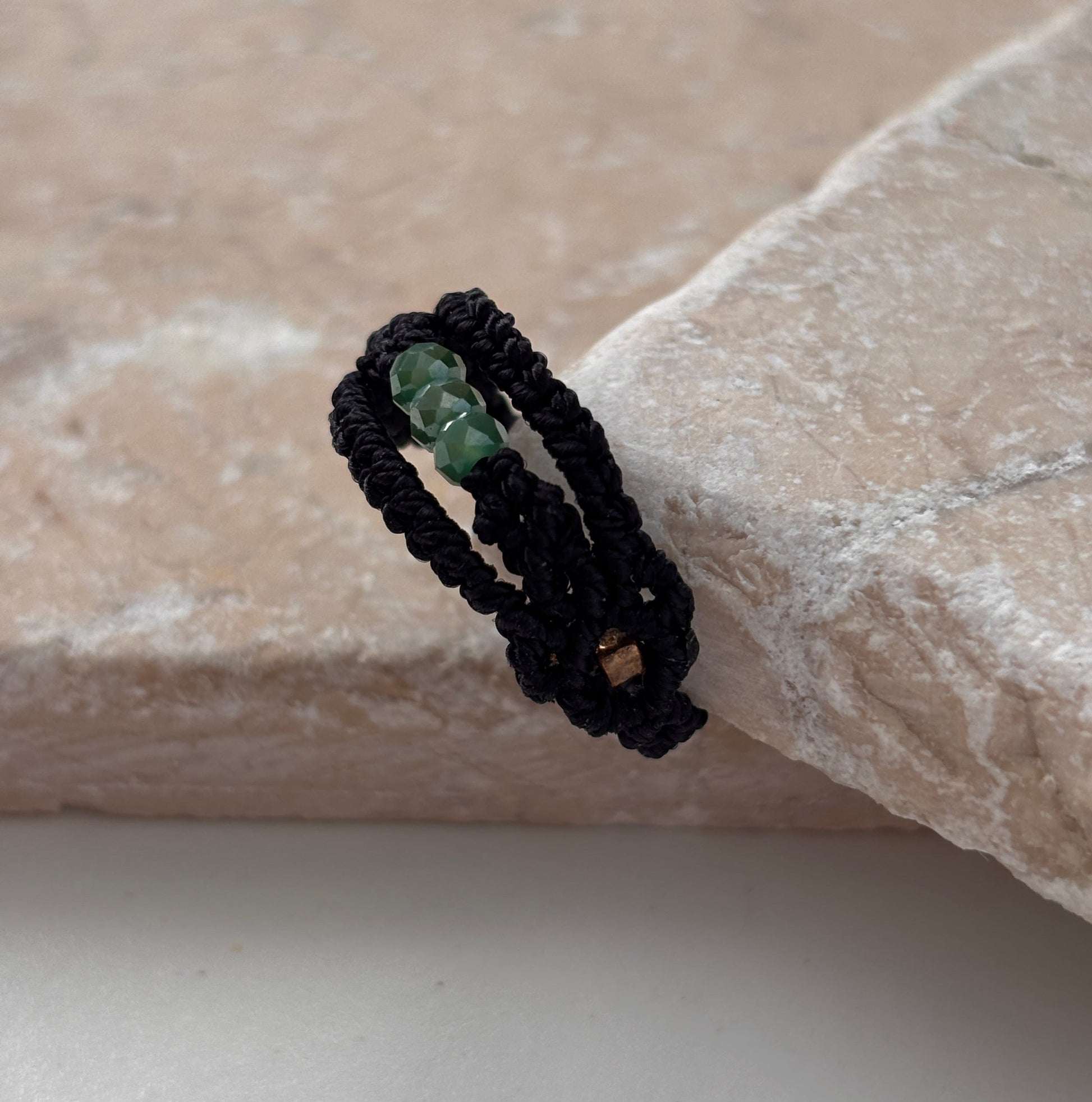 A side view of a handmade macrame band ring in black flat waxed nylon cord with a double-wrap braided design and three faceted mint green glass crystal beads with a square gold-tone seed bead accent visible on the underside, leaning against a pink stone surface against a white background,
