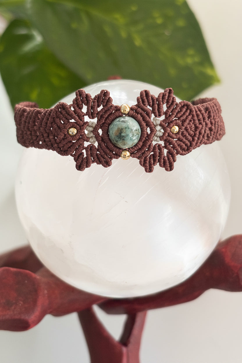 A handmade macrame bracelet in dark brown with a central round Indian Agate green stone and small brass beads, displayed on a clear crystal sphere stand.