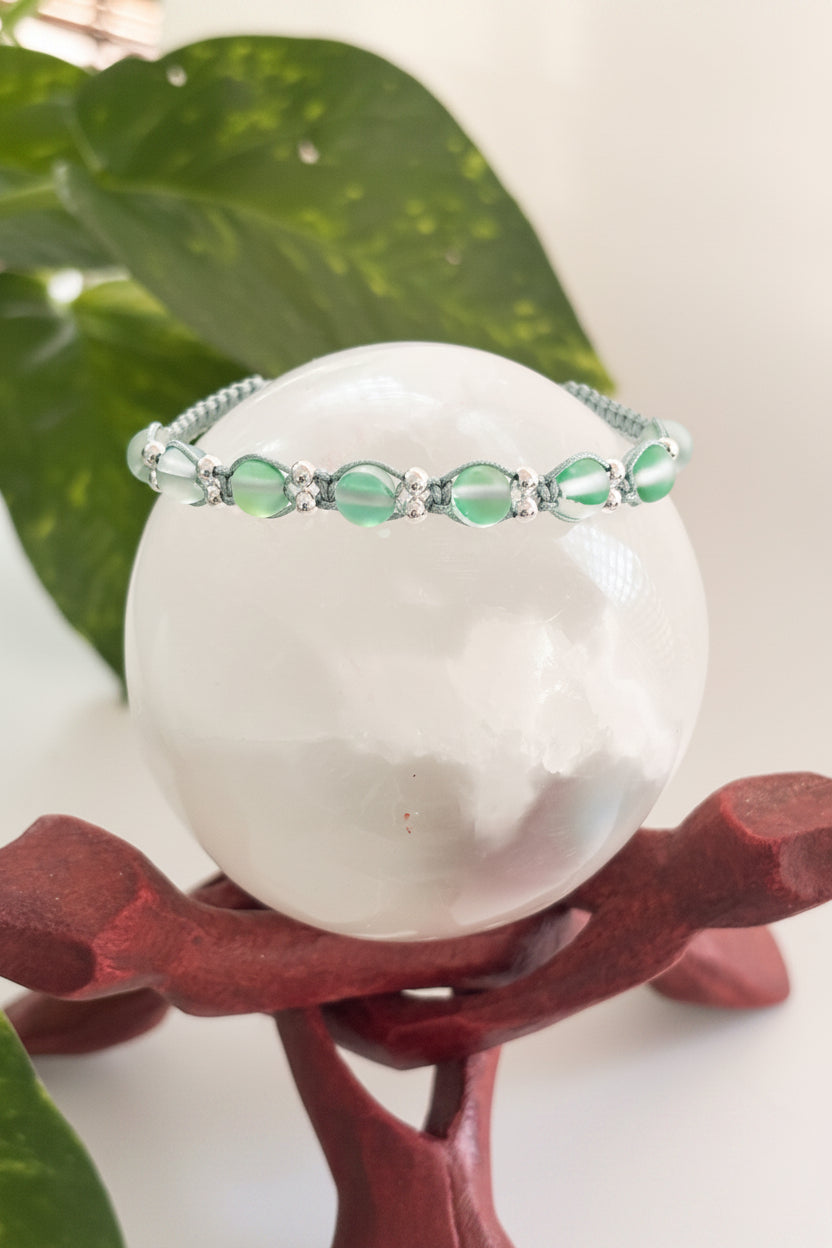 A sage green beaded macrame bracelet with silver-tone metal accent beads, displayed on a white crystal sphere atop a dark red stand with green leaves in the background.