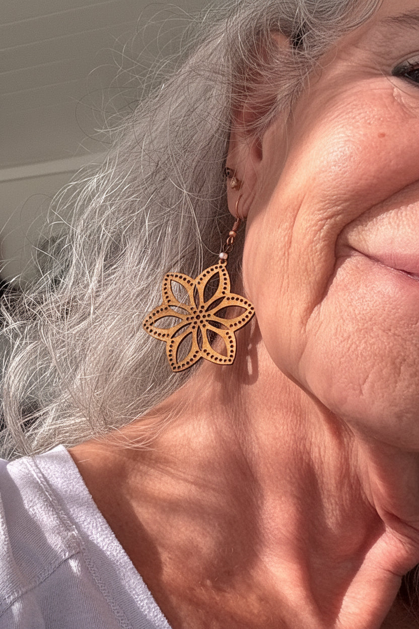 Handmade balsa wood flower earring with open petal silhouette and dotted border detail with a frosted peach seed bead accent on a copper fish hook wire, worn on a model with silver hair.