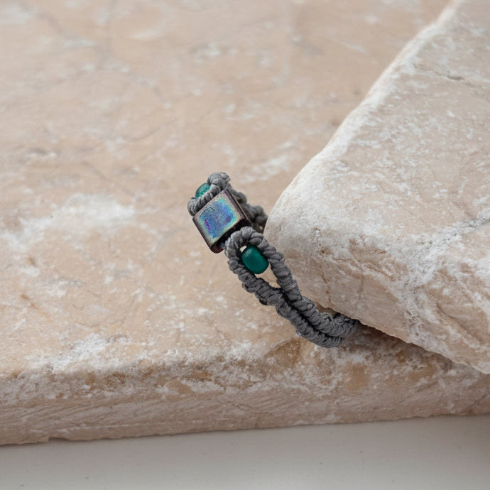 A side view of a handmade minimalist gray macrame ring with an iridescent blue-purple square Miyuki Tila bead at the center and two teal seed bead accents, resting on the edge of a pink stone surface against a white background