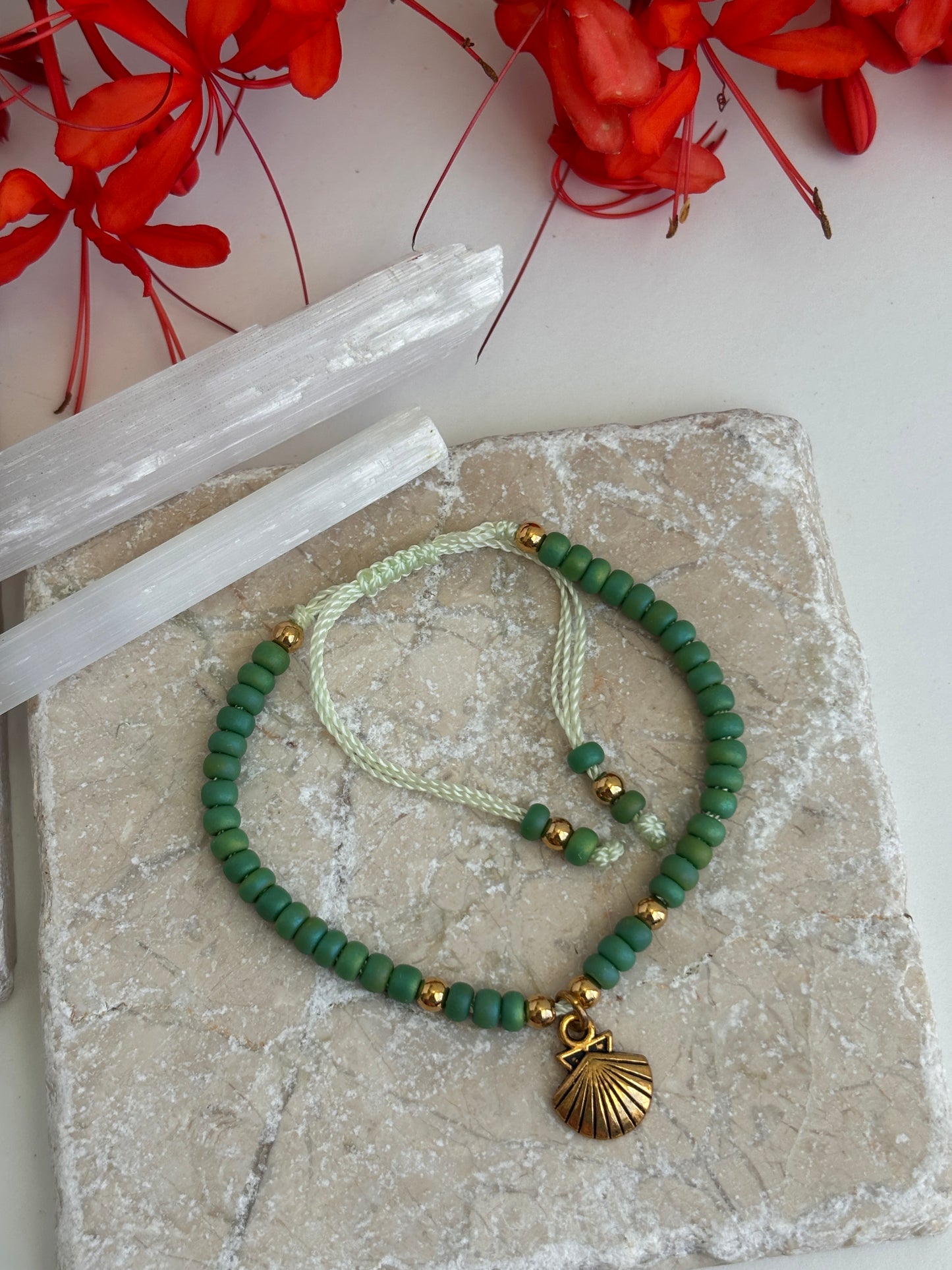 A beaded bracelet with green 6/0 seed beads, brass spacer beads, a mint green cord, and a brass shell pendant, displayed on a textured stone slab with red flowers and white crystal points in the background.