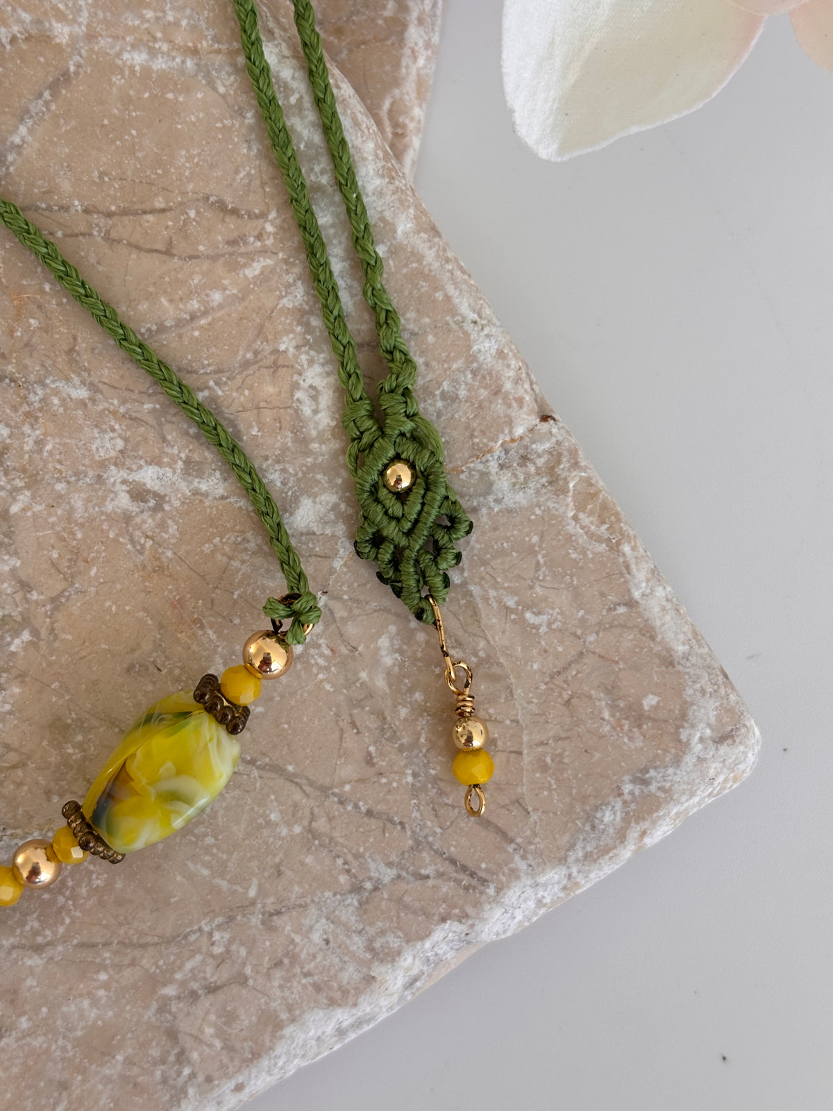 Close-up detail of handcrafted grass green macrame cord necklace showing decorative macrame slide closure with gold-tone bead, twisted rectangle chartreuse lampwork glass bead, 4mm round gold-tone beads, 3mm opaque yellow crystal, and brass rondelle spacers on travertine surface.