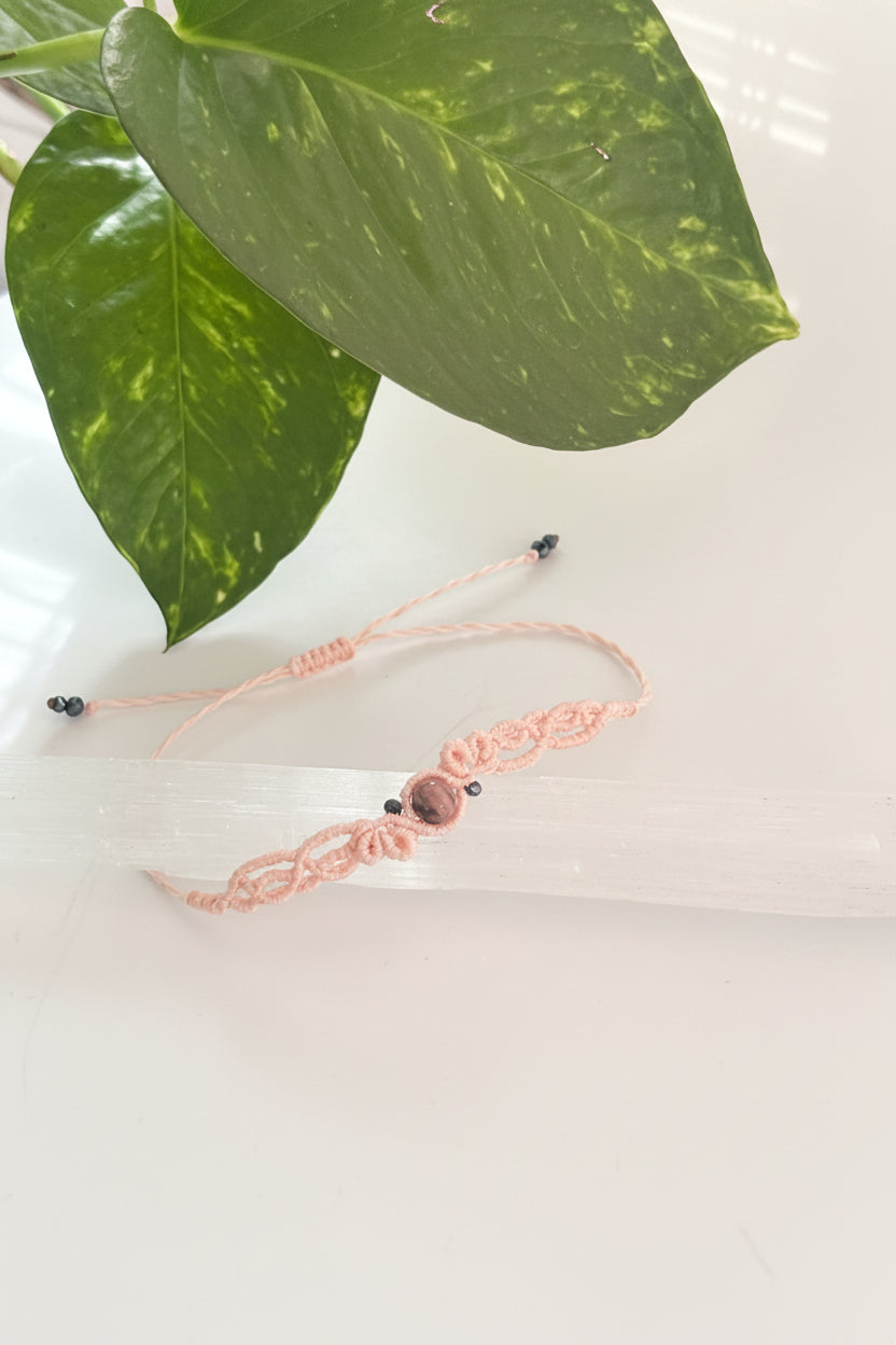 A light pink macrame bracelet with a central Rhodonite stone bead and black iridescent crystal beads, displayed on a clear acrylic block with green plant leaves in the background.