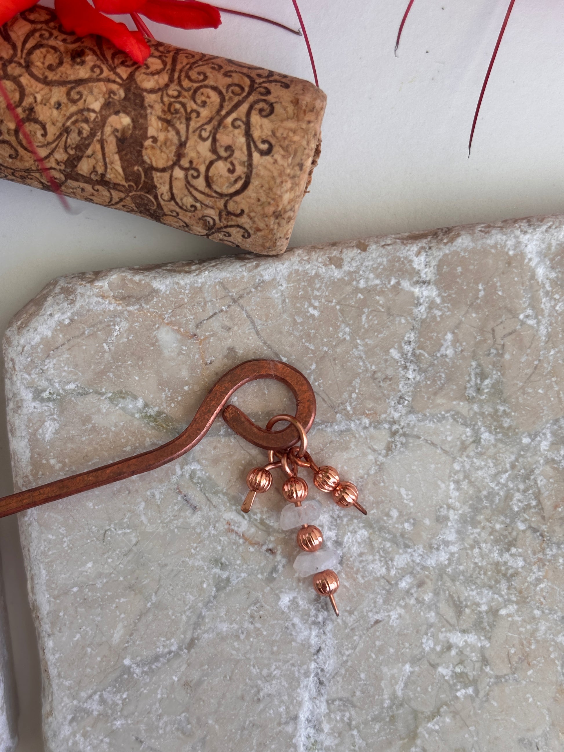 A hammered copper hair stick with three dangling copper beads and small moonstone accent beads, displayed on a textured stone slab with a cork and red leaves in the background.