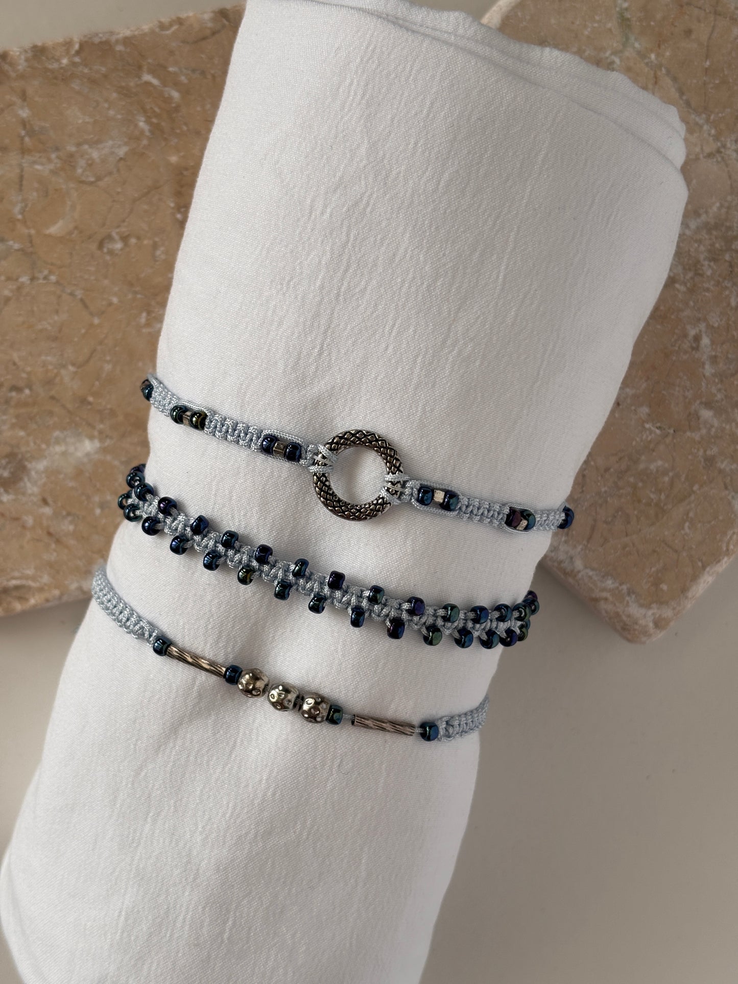 Three light blue macrame bracelets displayed on a white linen roll against a stone background — one with a silver-tone open ring centerpiece, one with dark metallic blue seed beads woven along the edges, and one with a silver-tone tube bead and silver-tone ball accent beads on light blue nylon cord.