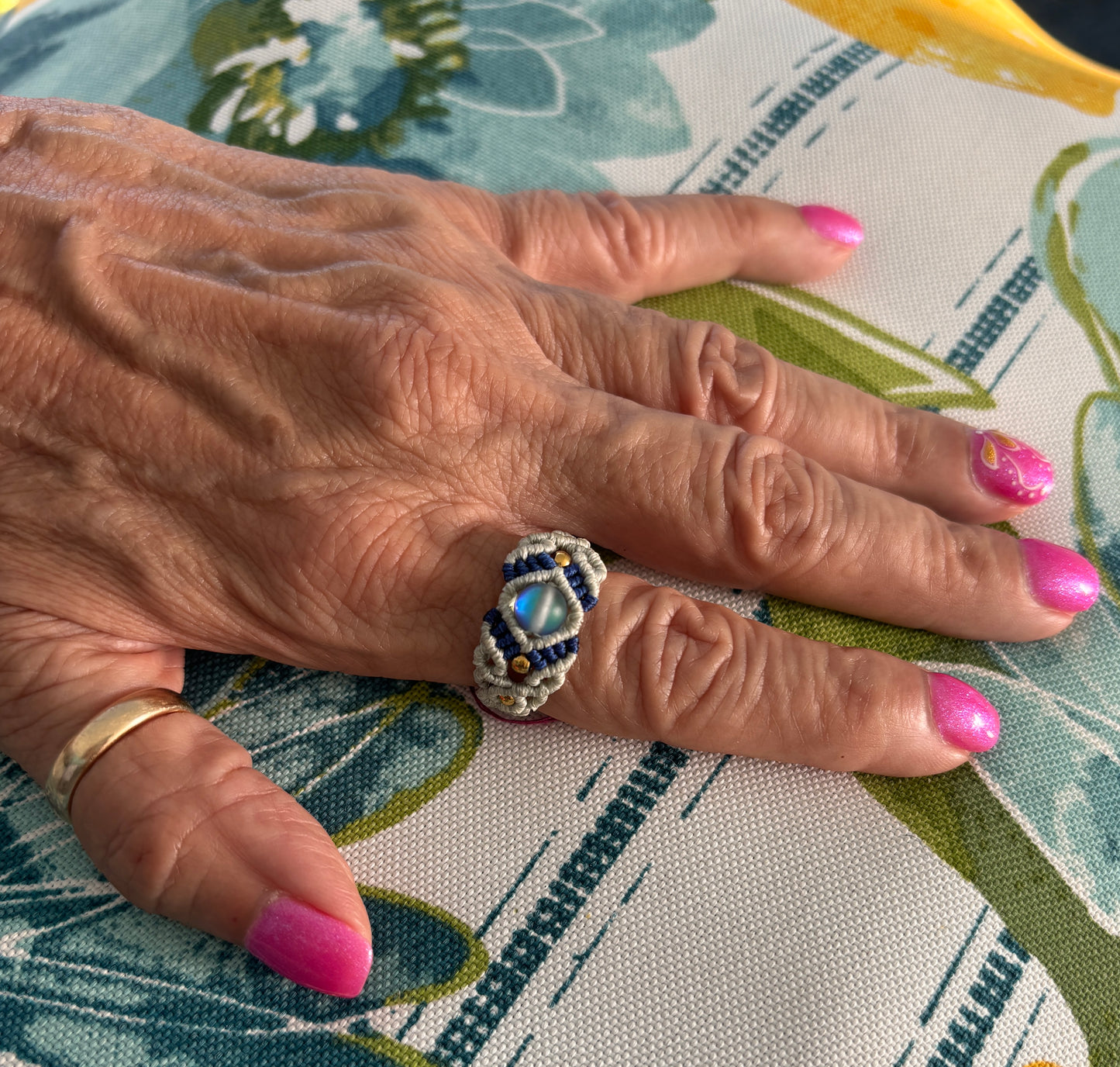 A handmade two-tone light gray and navy blue macrame cigar band ring with a shimmering iridescent mermaid glass bead and gold brass accent beads, worn on a finger with pink nails resting on a colorful floral fabric background.