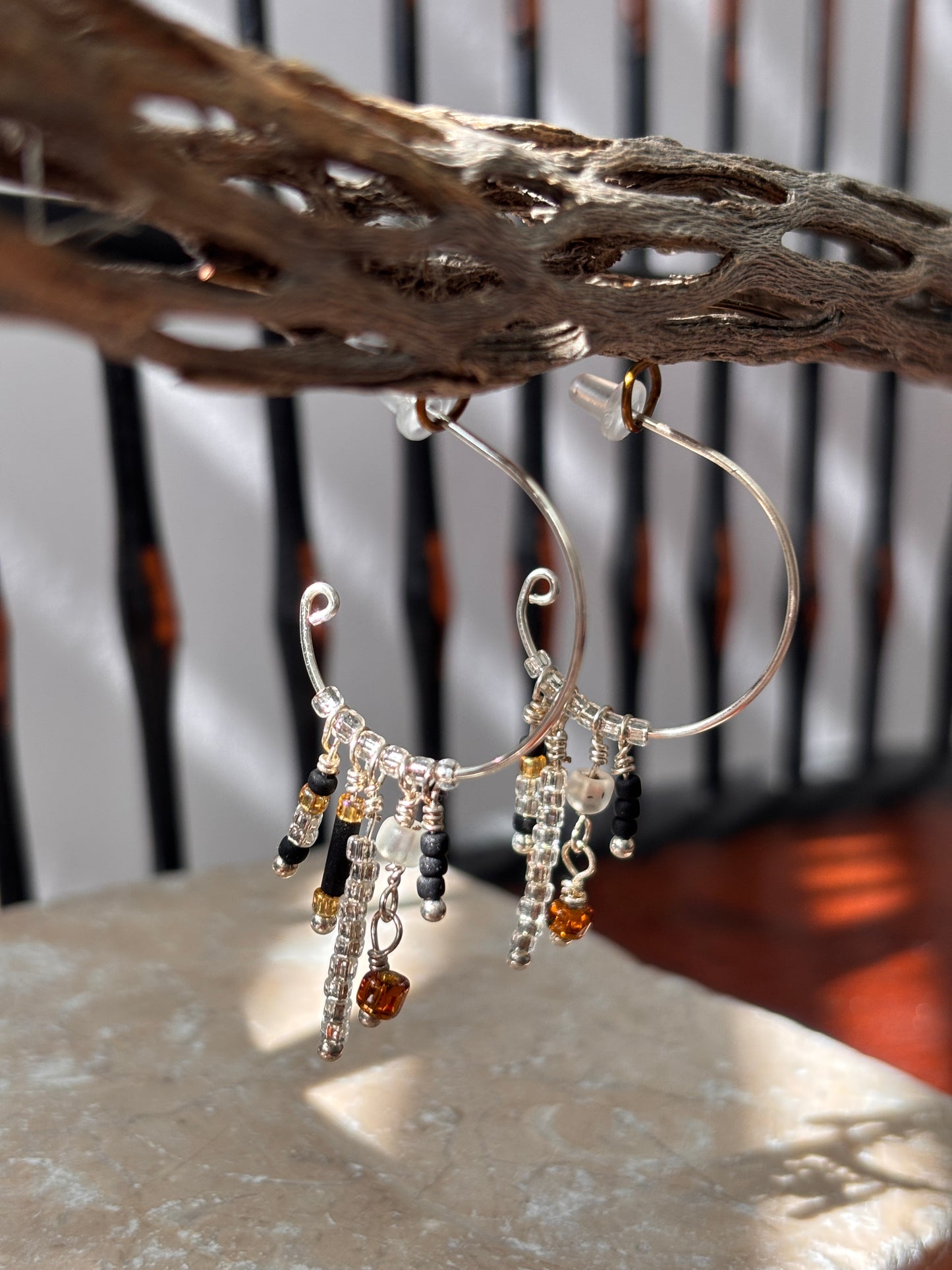 A pair of handcrafted silver-tone hoop earrings with a scroll-tip wire and five wire-wrapped dangle strands of black bugle beads, clear seed beads, frosted white disc beads, and an amber faceted cube bead drop, hanging from weathered driftwood in natural sunlight against a soft-focus outdoor background.