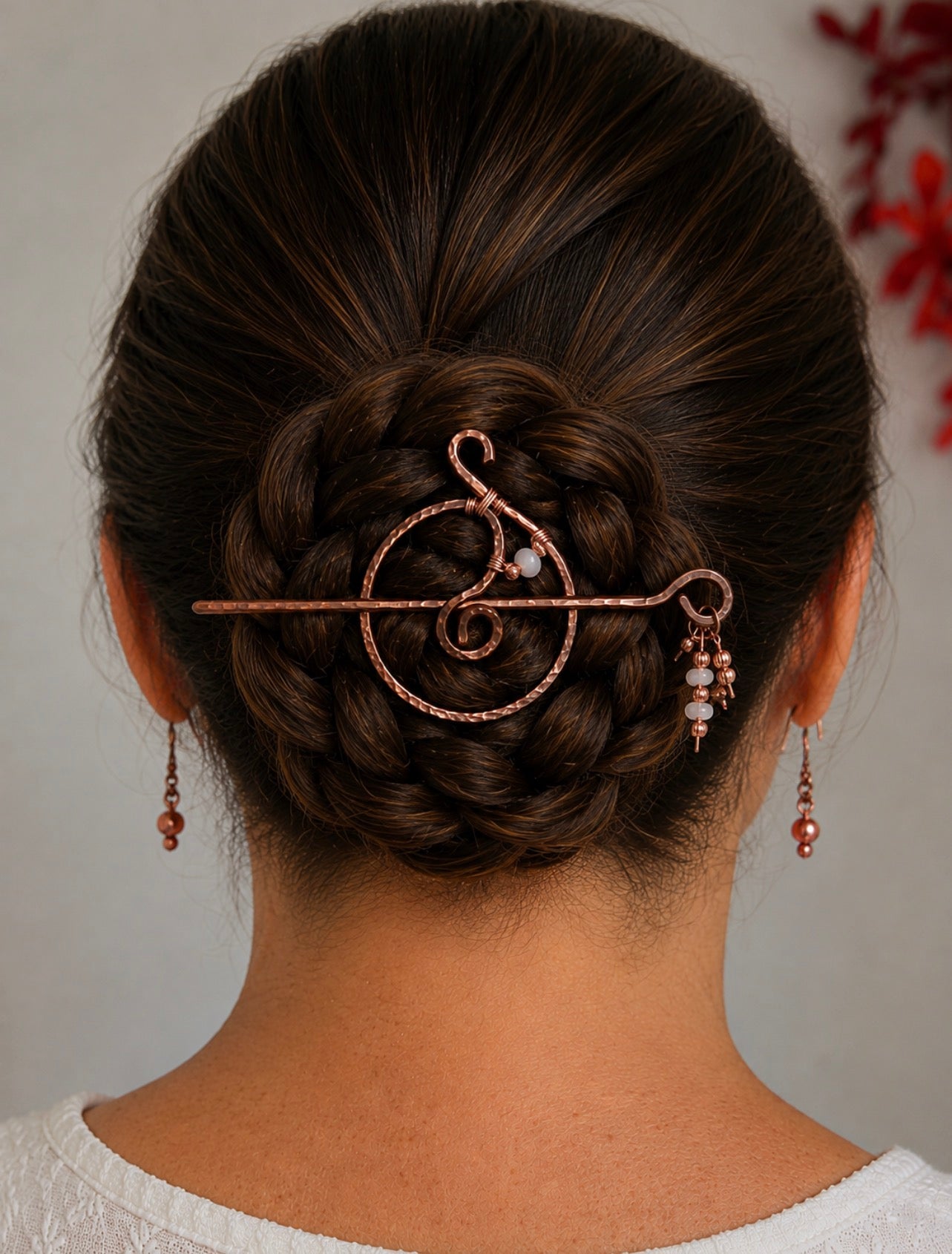 A woman viewed from the back wearing a hammered copper spiral hair pin accented with copper beads and moonstone beads in an updo bun, with matching dangling earrings, against a soft light gray background.