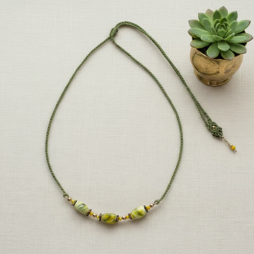 Handcrafted green macrame cord necklace with twisted rectangle chartreuse lampwork glass beads on a brass wire bar and small brass spacer beads, laid flat on an ivory linen background.