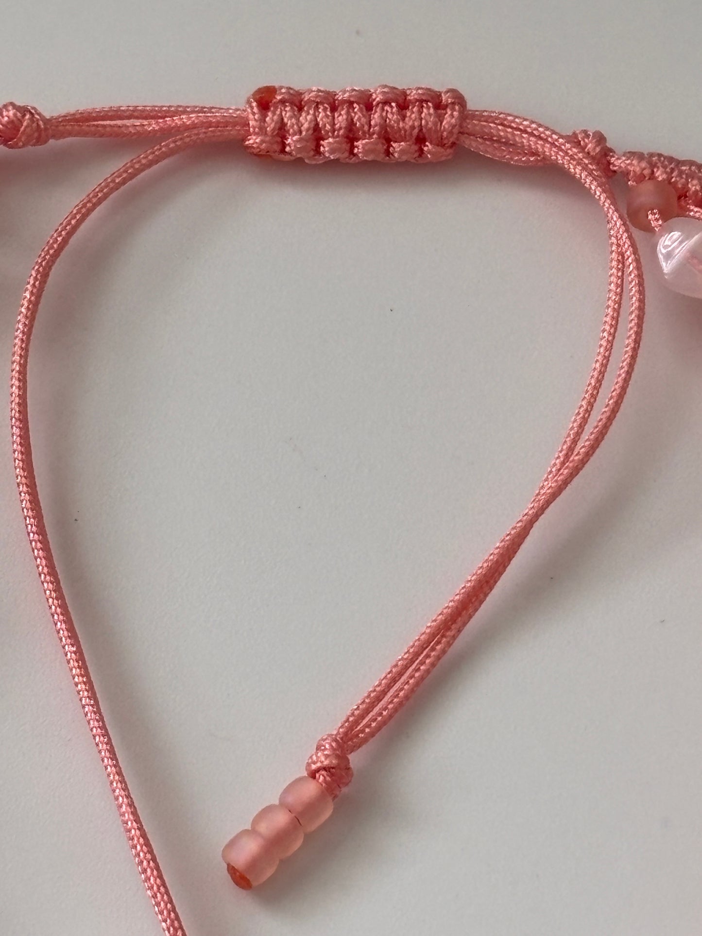 A close-up of the adjustable slide closure on a handmade light coral macrame anklet, showing the knotted slide mechanism, white opal bead and matte peach Myuki seed beads on a white background