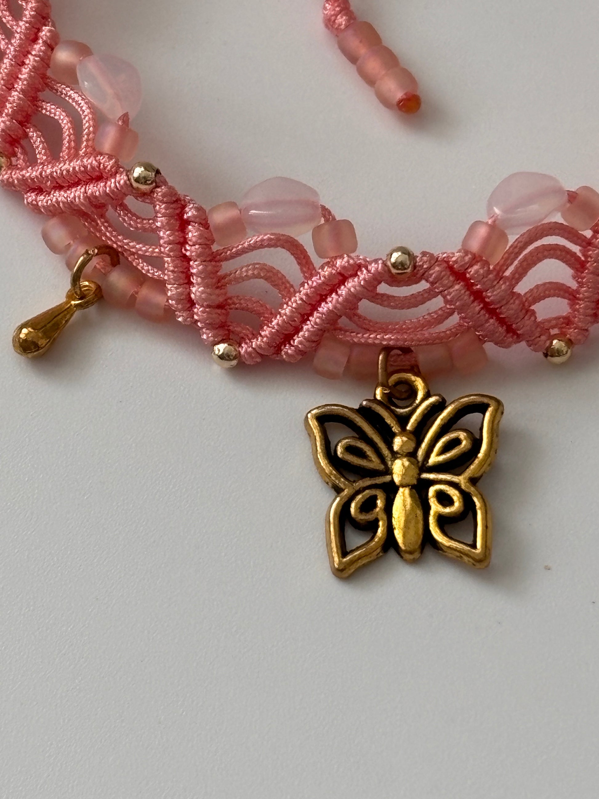 A close-up of a handmade light coral macrame anklet showing the double zig-zag knotwork, opal white Preciosa beads, matte peach Miyuki seed beads round brass beads, a gold-tone butterfly charm, and a gold-tone water drop charm on a white background.