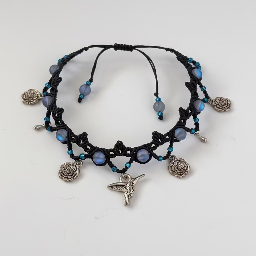 A black macrame-style beaded anklet with small blue seed beads, frosted iridescent blue mermaid glass beads, silver-toned flower, hummingbird and water drop charms, displayed on a white background.