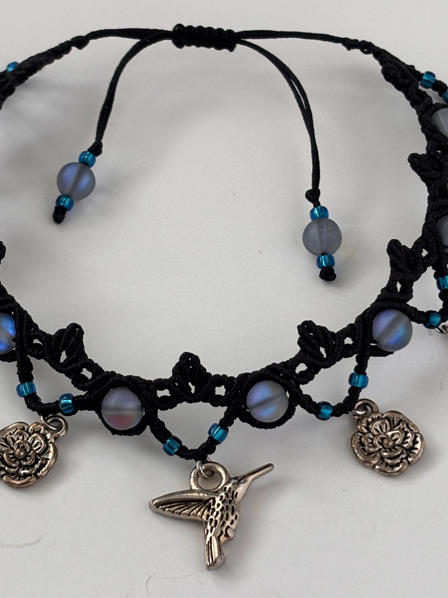 Black macrame anklet with frosted iridescent blue mermaid glass beads, small blue seed beads, and silver-toned charms including a hummingbird, flower, and water drop displayed on a white background.