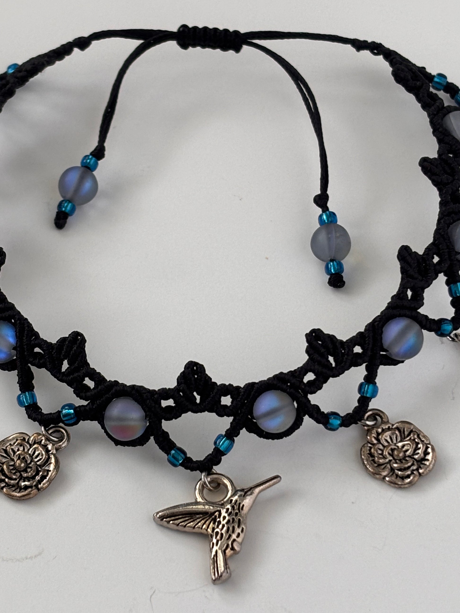 Black macrame anklet with frosted iridescent blue mermaid glass beads, small blue seed beads, and silver-toned charms including a hummingbird, flower, and water drop displayed on a white background.