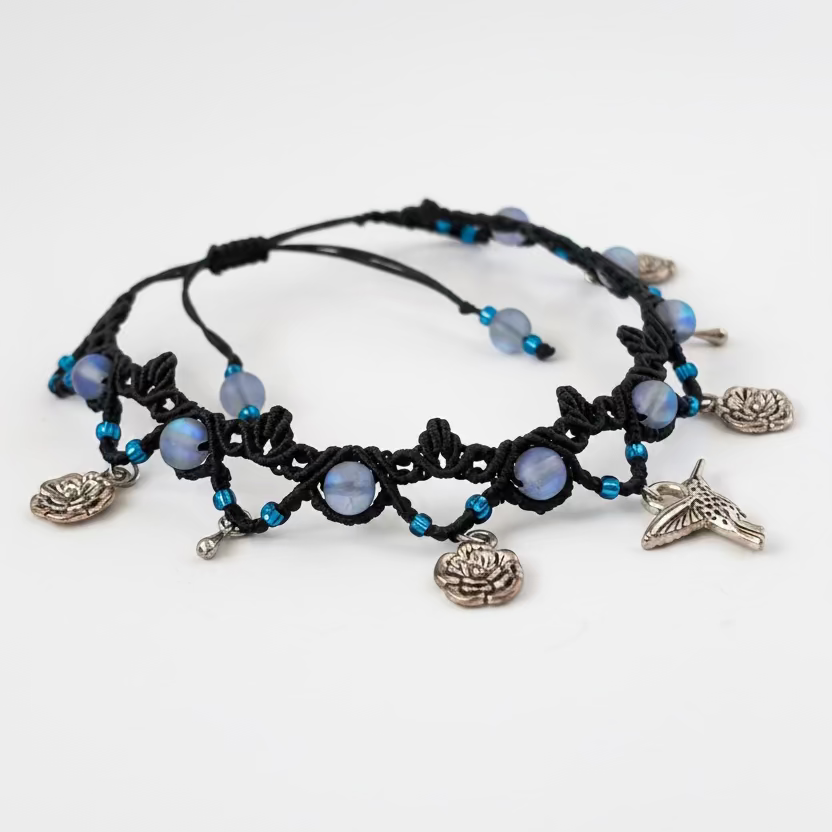 Black macrame anklet with frosted  iridescent blue mermaid glass beads, blue seed beads, and silver-toned charms including a hummingbird, flower and water drop charm, displayed on a whit background.