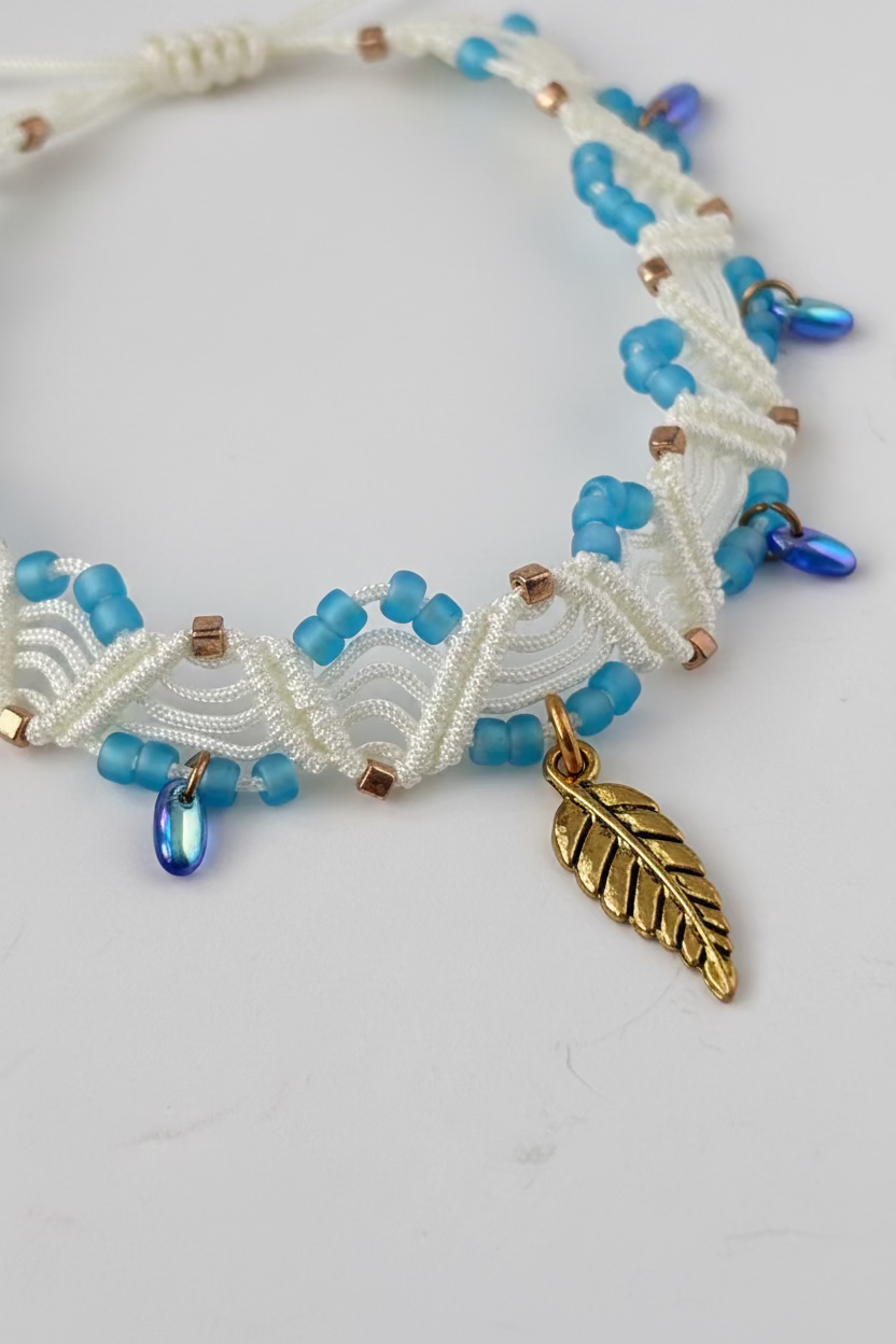A close-up of a handmade ivory macrame anklet showing the double zig-zag knotwork with turquoise matte seed beads, gold Miyuki square seed beads, iridescent blue Czech glass water drop beads, and a gold-tone leaf charm on a white background.