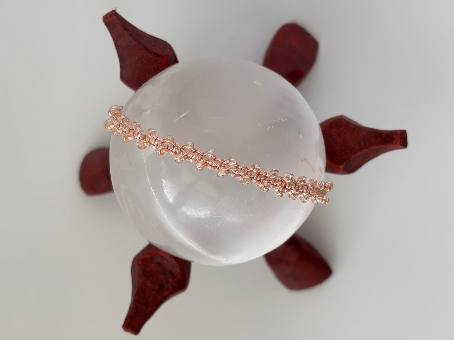 A delicate light peach beaded macrame bracelet displayed horizontally across a smooth, translucent white crystal sphere, which is supported by a dark reddish-brown decorative stand on a light gray background.