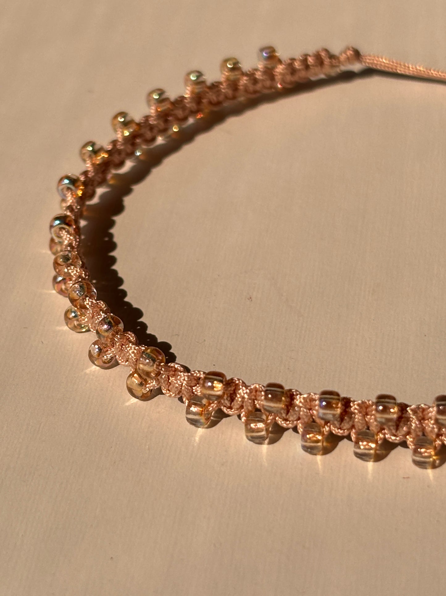 A close-up of a lightpeach beaded macrame bracelet with yellow irridescent seed beads and a textured woven pattern, displayed on a neutral beige background.