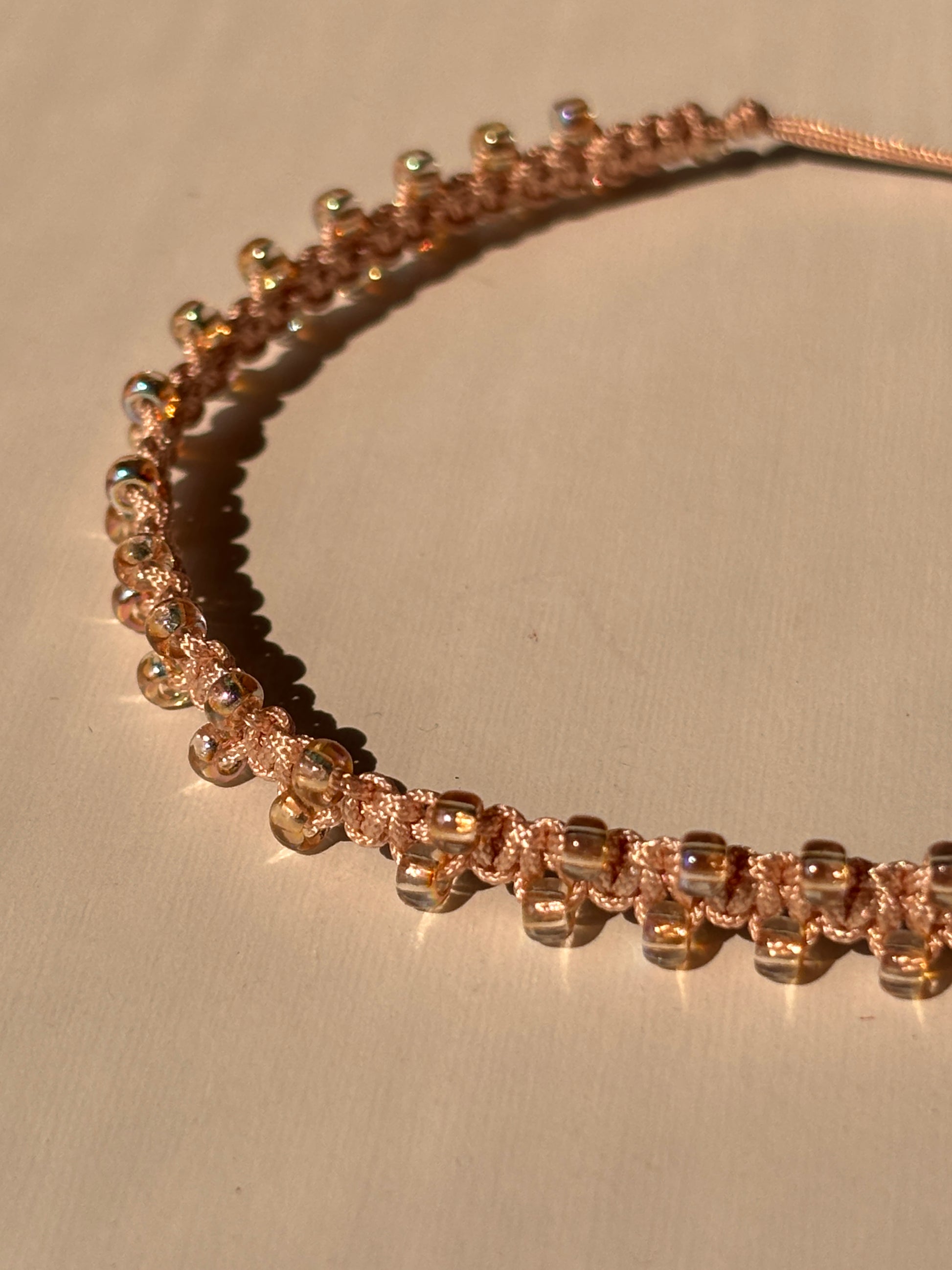 A close-up of a lightpeach beaded macrame bracelet with yellow irridescent seed beads and a textured woven pattern, displayed on a neutral beige background.