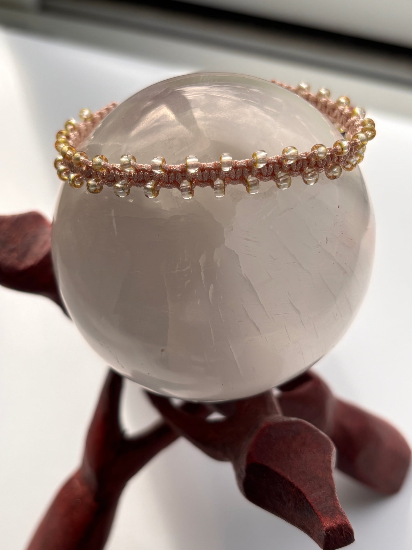 A translucent pale gray stone sphere displayed on a dark reddish-brown stand, with a thin beaded macrame bracelet draped over one side in light peach and copper edged seed beads.