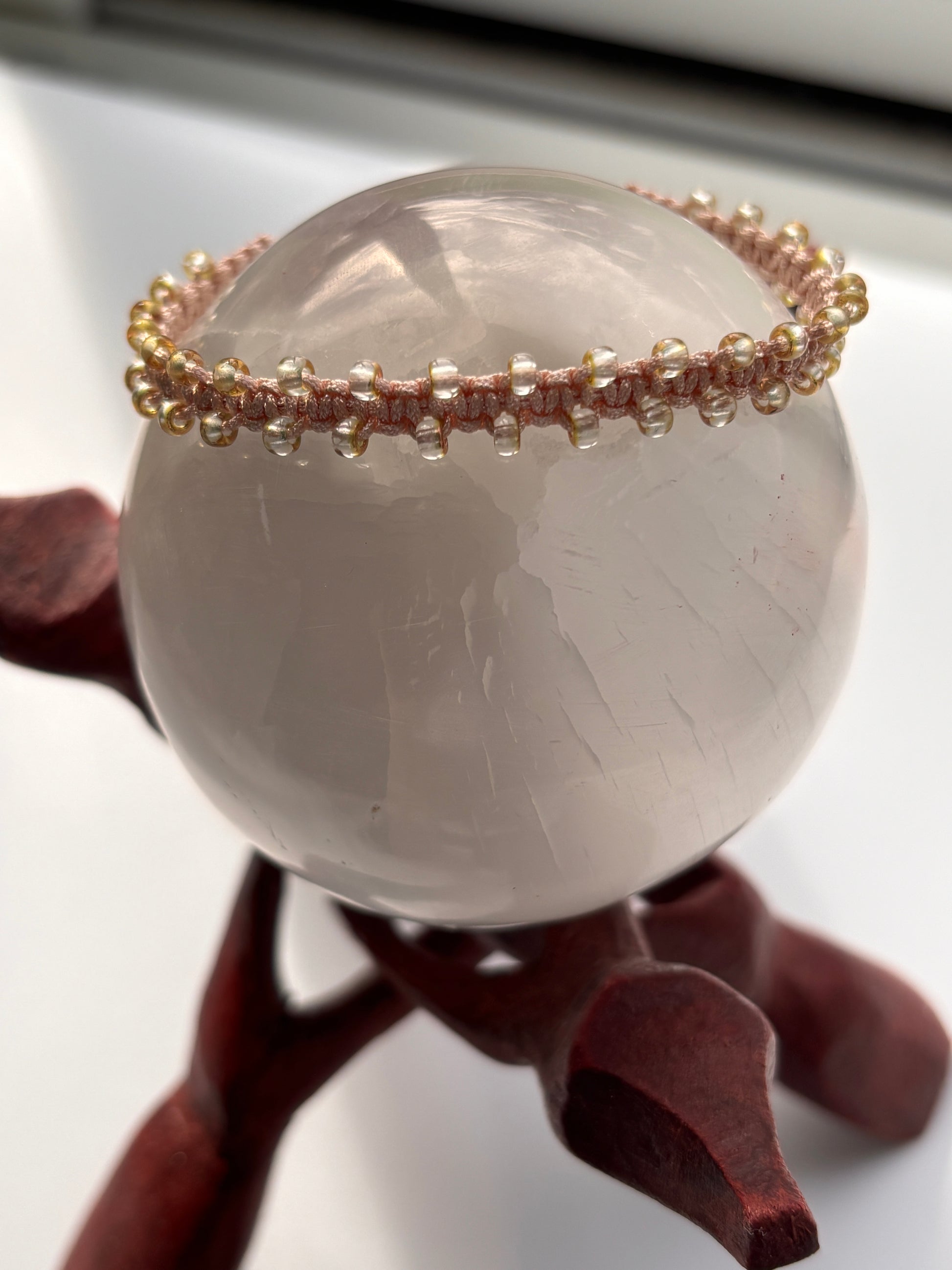 A translucent pale gray stone sphere displayed on a dark reddish-brown stand, with a thin beaded macrame bracelet draped over one side in light peach and copper edged seed beads.