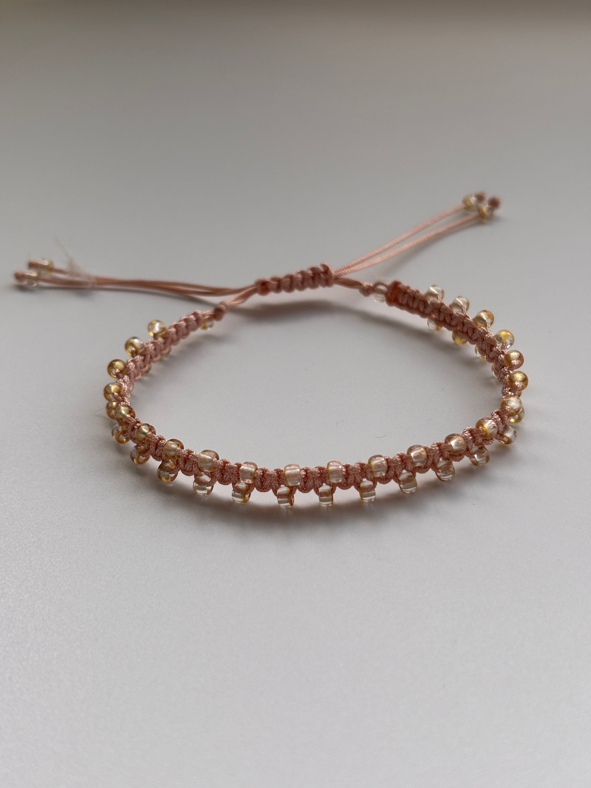 A handmade beaded macrame bracelet with metallic edged seed beads, featuring an adjustable cord closure, displayed against a plain light gray background.