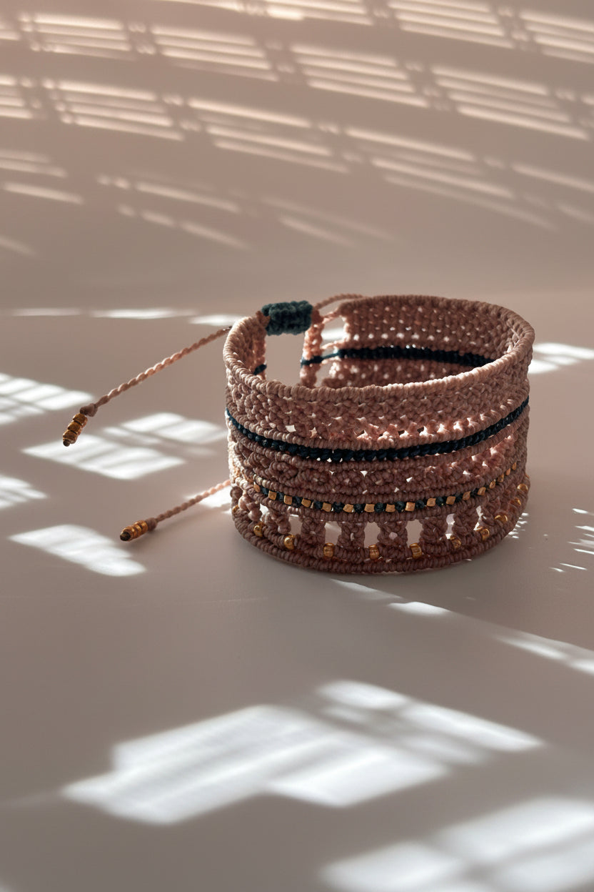 A macrame cuff bracelet made with warm taupe-brown color cord and accented with teal cord and metallic cold-tone beads, photographed against a white wall with dramatic window shadow patterns.