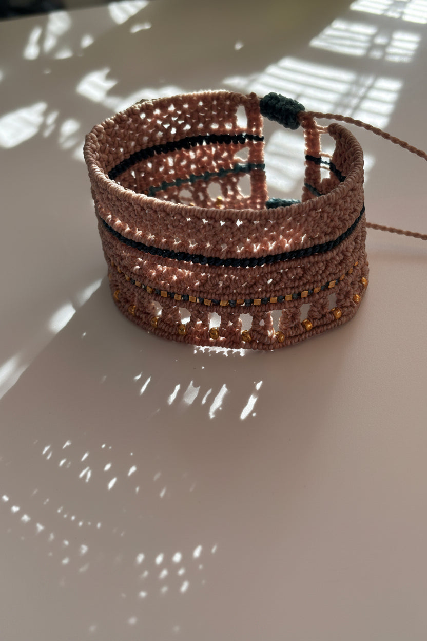 A macrame cuff bracelet made with warm taupe-brown color cord accented with teal cord and gold-tone metal beads, photographed against a light wall with dappled sunlight and shadows.