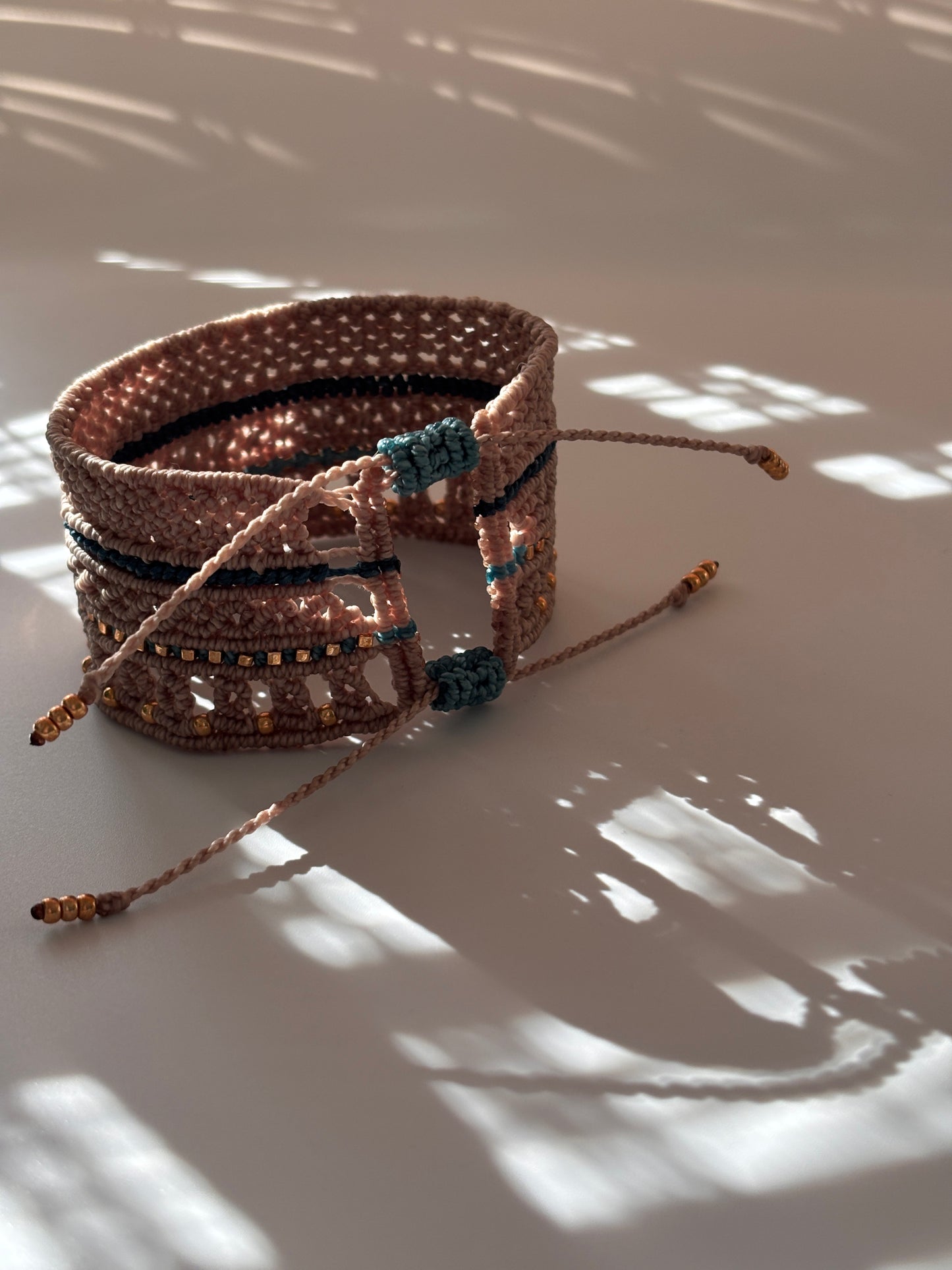 A macrame cuff bracelet made with warm taupe-brown color cord with teal accents and gold-tone metal bead details, photographed against a white wall with dappled sunlight and shadows.
