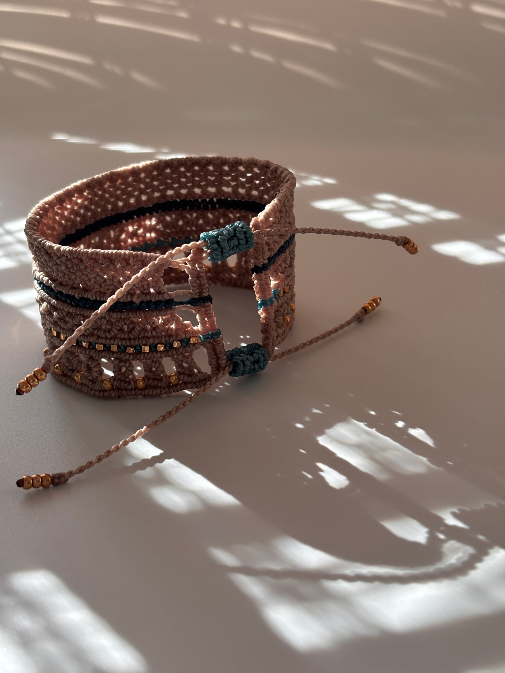 A macrame cuff bracelet made with warm taupe-brown color cord with teal accents and gold-tone metal bead details, photographed against a white wall with dappled sunlight and shadows.