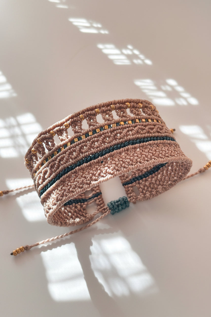 A thick macrame cuff bracelet in a warm taupe-brown color, decorated with teal cord and small gold bead accents, hanging against a neutral wall with soft window shadow patterns.