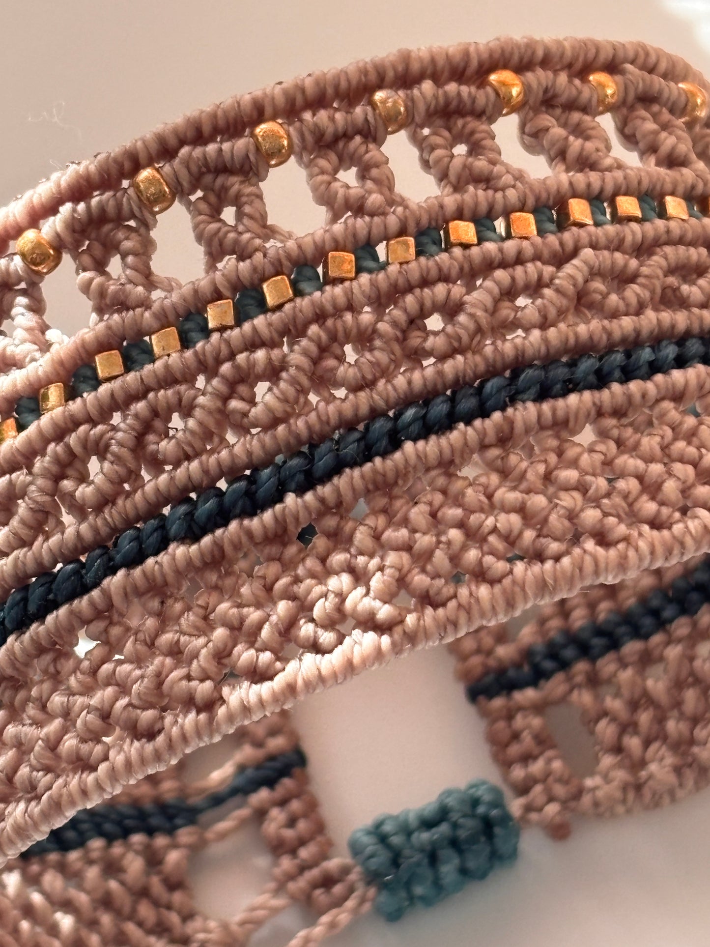 A close-up of a handmade macrame bracelet featuring textured beige cords, teal braided stripes, and small metallic gold beads.