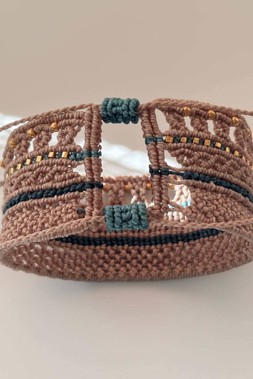 A close-up of a macrame cuff bracelet  with a taupe-brown base, dark teal stripes, and small gold bead accents, photographed against a plain light background.