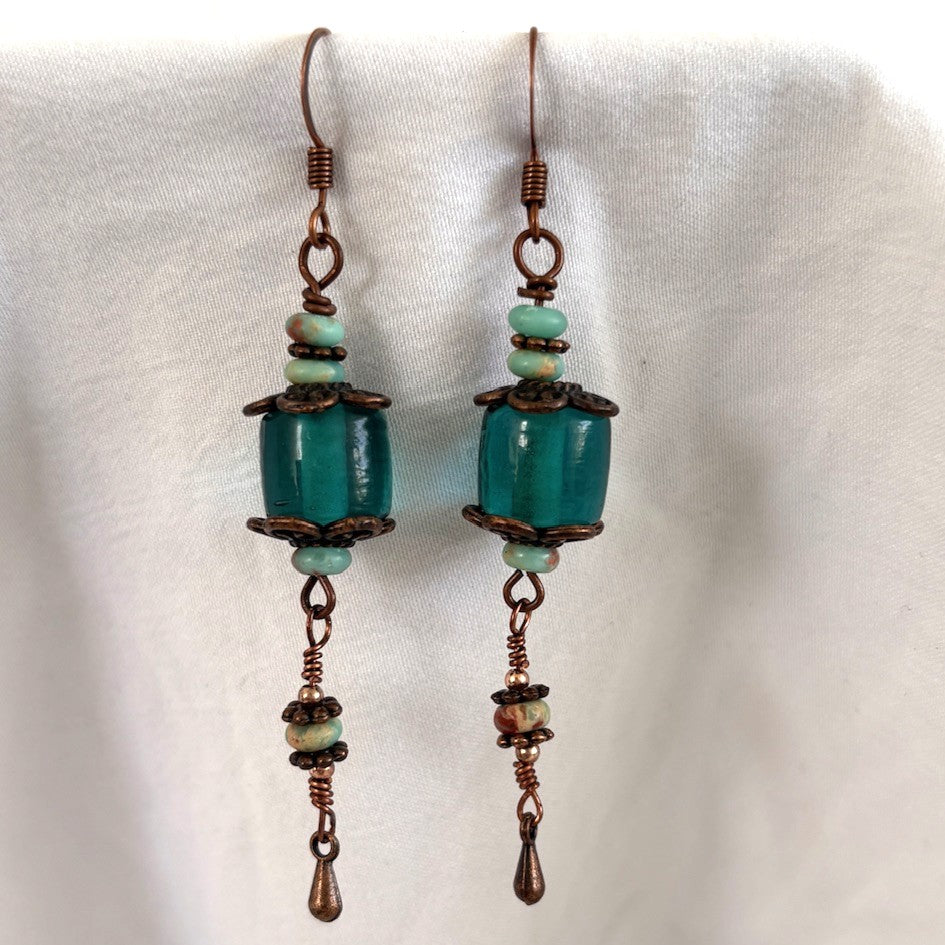 Close-up of handmade teal dyed glass drop earrings showing copper filigree caps, abacus-shaped blue sea sediment beads, 10mm × 10mm teal glass bead, and wire-wrapped copper waterdrop charm, displayed against white fabric.