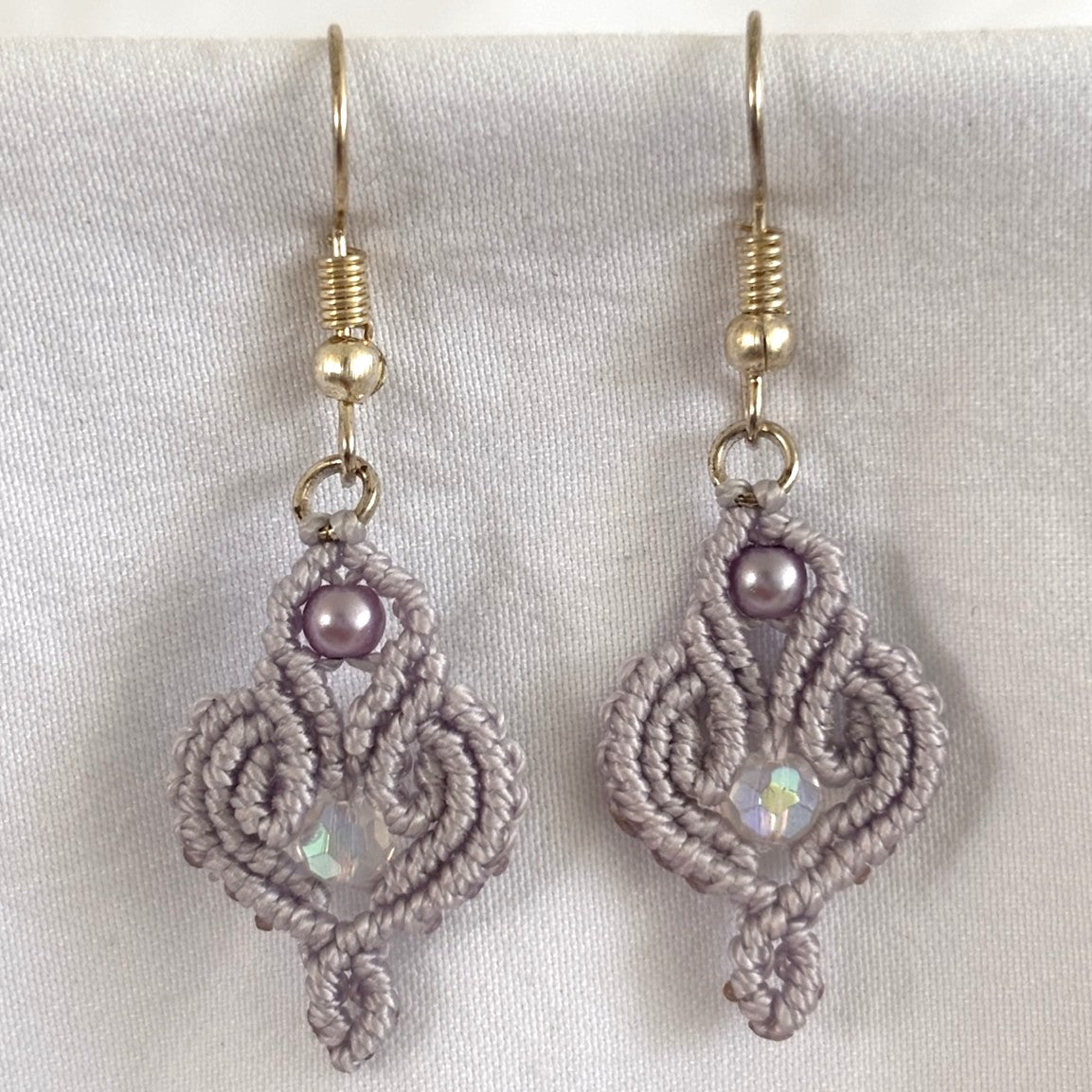 Close-up of handmade lavender macrame scroll drop earrings showing ornate knotwork scrolls, 3mm lavender glass pearl accent, and 4mm faceted iridescent crystal center, displayed against white fabric.