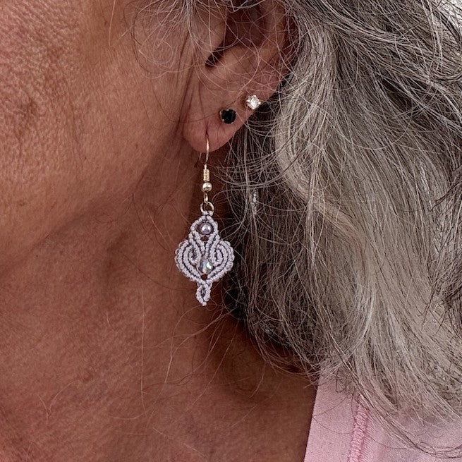 Handmade lavender macrame scroll drop earring with glass pearl and iridescent crystal center shown worn on model with silver hair.