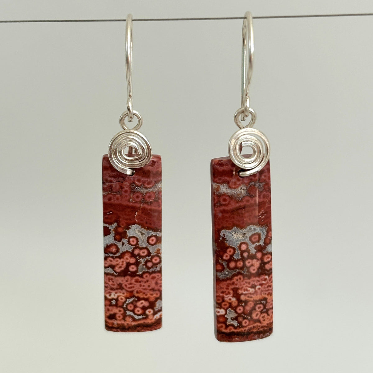 Handmade brecciated jasper earrings with silver-tone handcrafted swirl bail and hammered ear wires, hanging against a neutral gray background showing full stone and bail detail