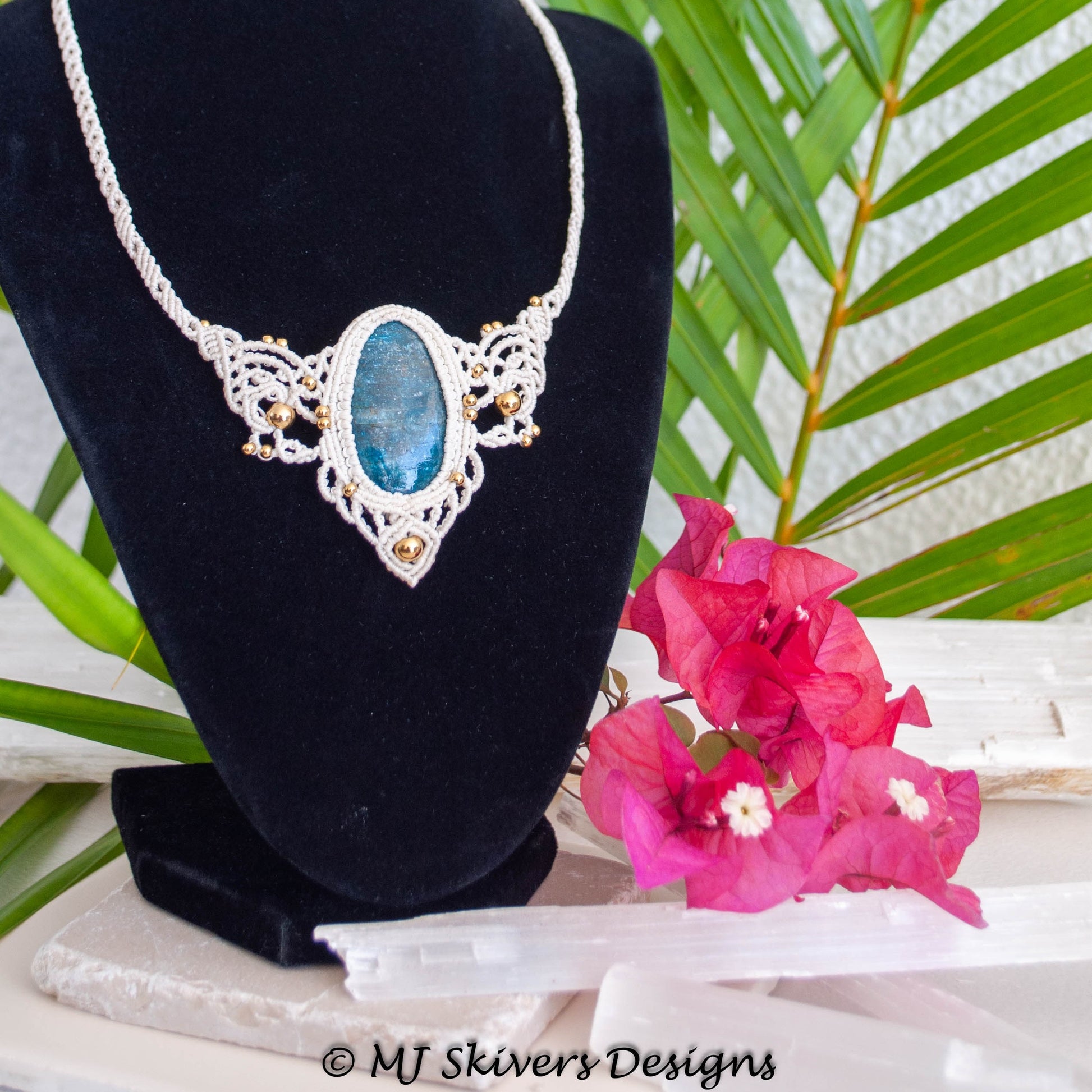 A silver-toned macrame necklace with an oval blue gemstone pendant, displayed on a black velvet jewelry bust. The bust is styled with bright pink bougainvillea flowers and green palm leaves in the background.