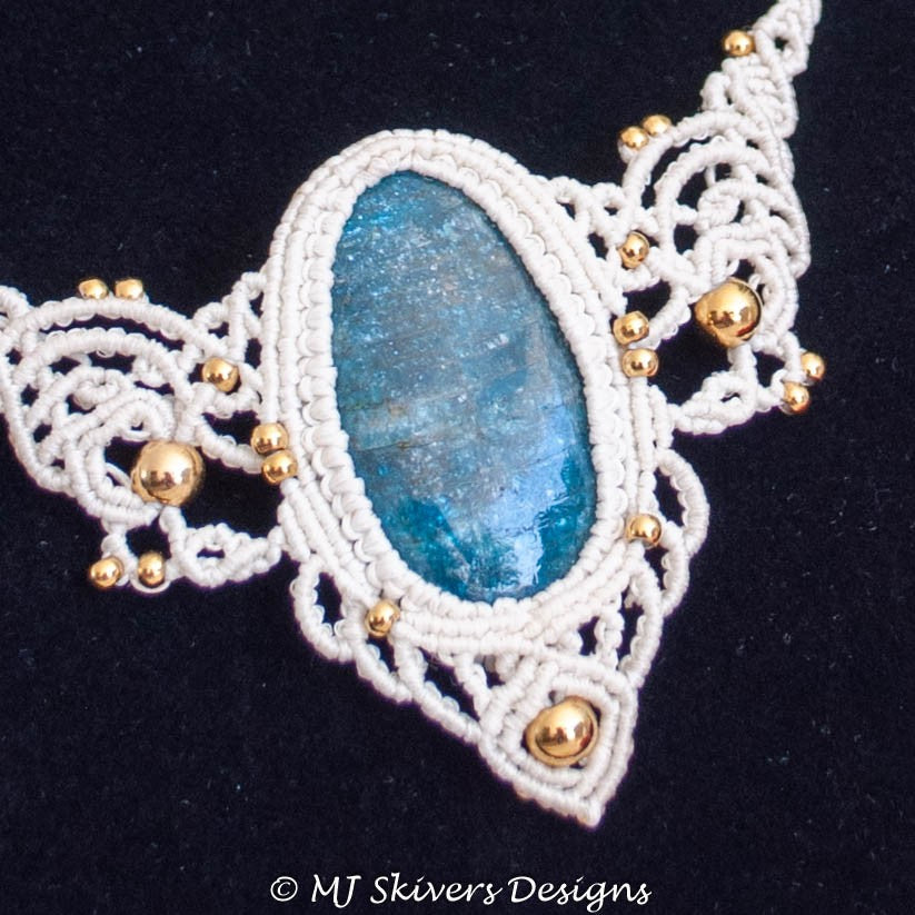 A close-up of a handmade macrame necklace featuring a central oval blue stone, intricate white woven cord, and small gold beads, set against a black background.