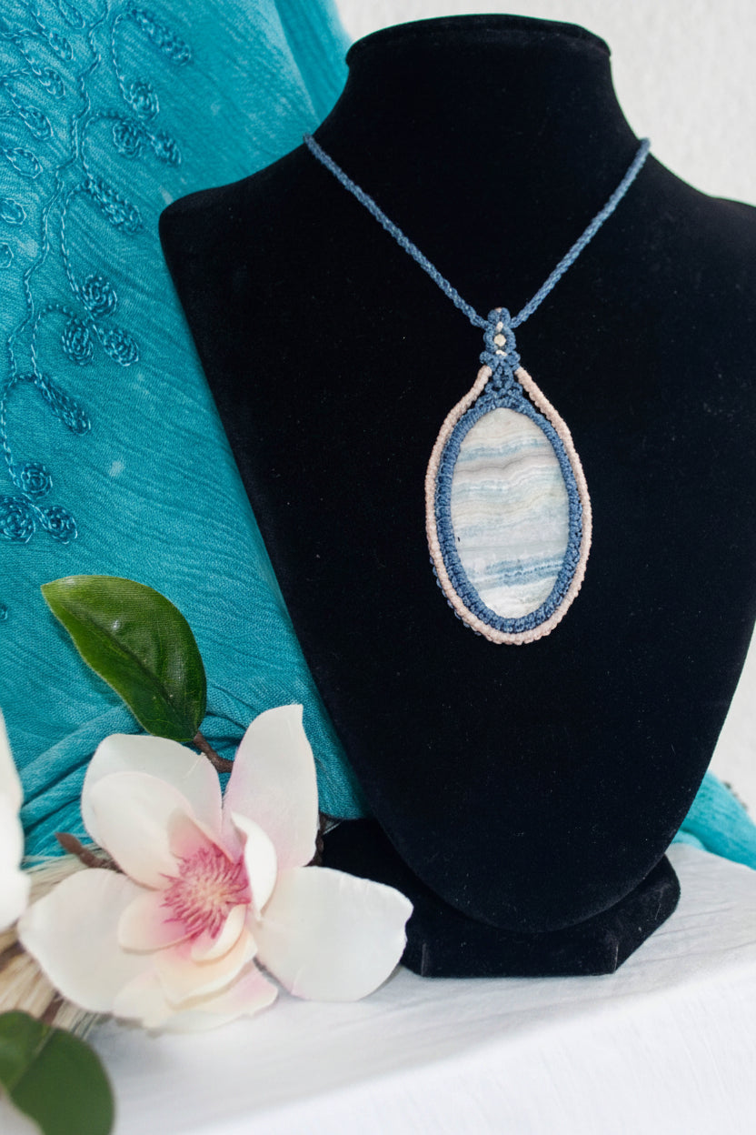 A handmade marame necklace with a Blue Scheelite oval cabochon pendant, displayed on a black velvet jewelry bust. The necklace features a blue macrame frame around the stone and an ivory macrame cord around the blue.