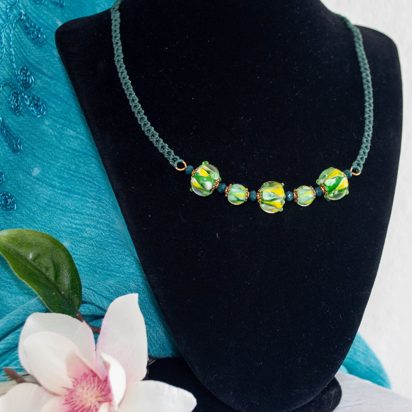 A green beaded necklace displayed on a black velvet jewelry bust, featuring five textured beads with a green and yellow marbled pattern. A turquoise fabric backdrop and a white flower are visible in the background.