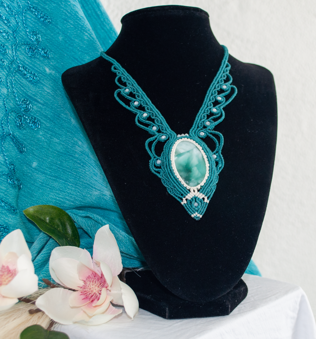 A teal and wh itemacrame necklace with a central Hemimorphite  stone, displayed on a black velvet jewelry bust. The bust is styled with a teal fabric drape and white orchids on a white tablecloth.
