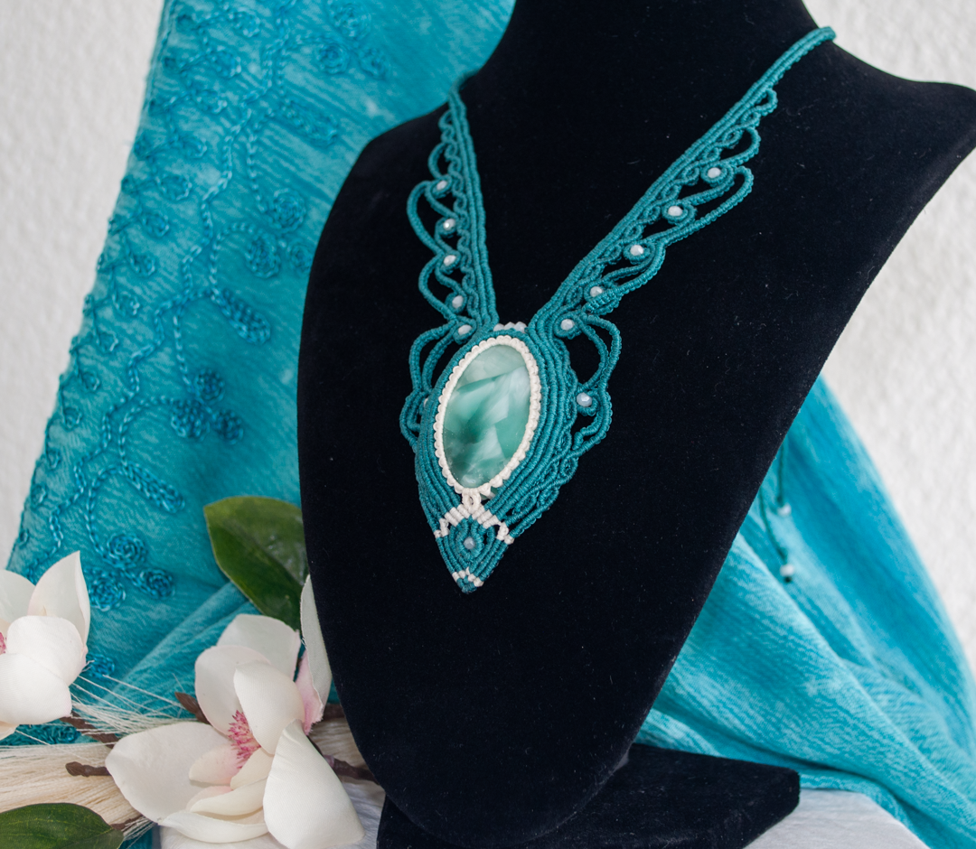 A teal and white beaded macrame necklace with a large oval Hemimorphite stone pendant, displayed on a black velvet jewelry bust with a turquoise lace fabric backdrop and white flowers in the foreground.