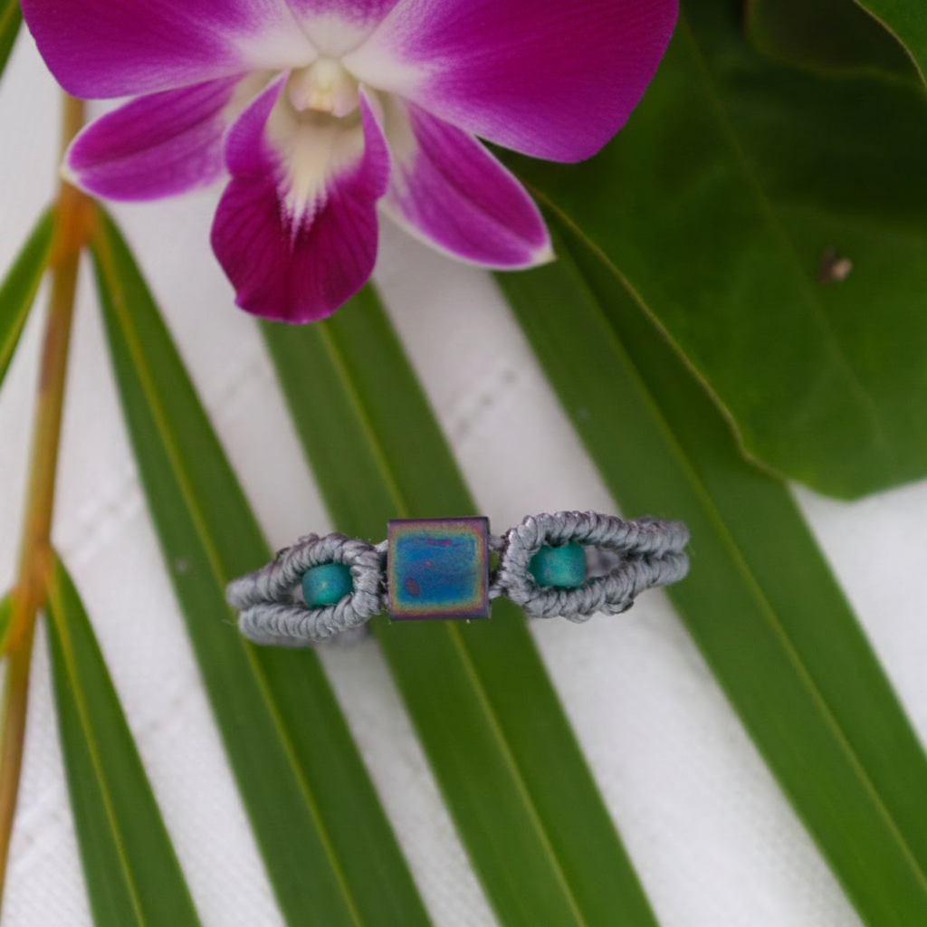 A handmade minimalist gray macrame ring with an iridescent square Miyuki Tila bead at the center and vibrant teal seed beads, displayed flat on a tropical green leaf with a magenta flower and marble surface in the background.