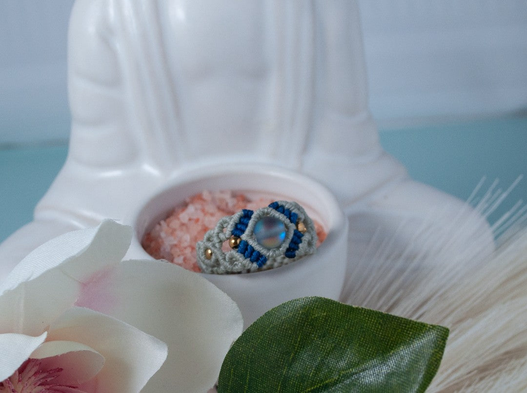 A handmade two-tone light gray and navy blue macrame cigar band ring with a frosted iridescent mermaid glass bead and gold brass accent beads, displayed in a white ceramic Buddha dish with pink Himalayan salt, a white flower petal, and a green leaf, against a soft blue background.