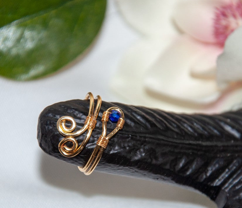 A handcrafted gold-tone wire ring with a double spiral flourish and a 3mm deep blue faceted crystal bead wire-wrapped at the base, displayed on a carved black decorative hand prop with a white magnolia flower and green leaf in the background