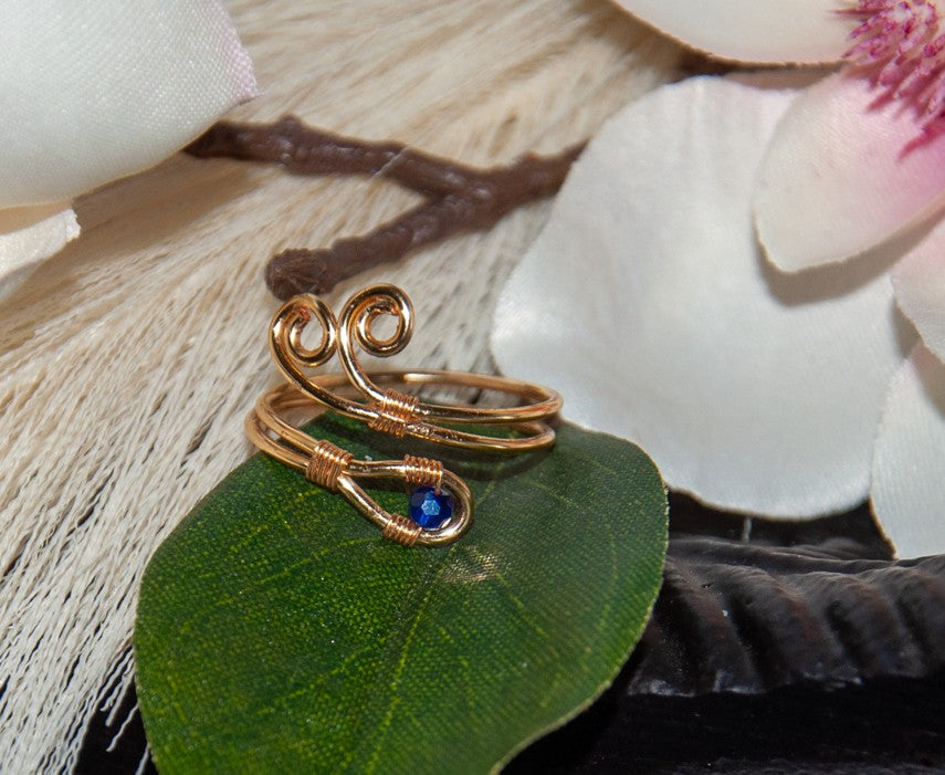 A handcrafted gold-tone wire ring with a double spiral flourish and a 3mm deep blue faceted crystal bead wire-wrapped at the base, resting on a green leaf with white and pink magnolia flowers and driftwood in the background