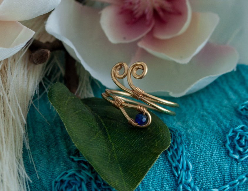 A handcrafted gold-tone wire ring with a double spiral flourish and a 3mm deep blue faceted crystal bead wire-wrapped at the base, resting on a green leaf against a teal embossed surface with a white and pink magnolia flower in the background